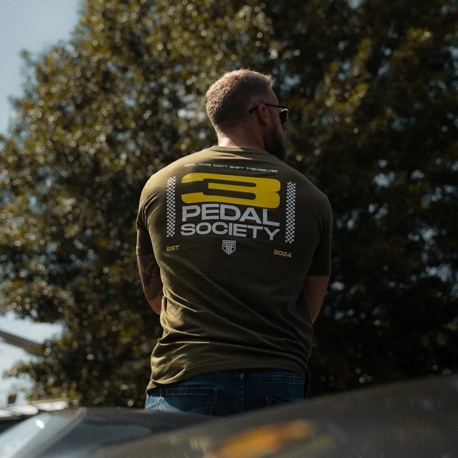 Man wearing a '3 Pedal Society' t-shirt standing outdoors with trees in the background