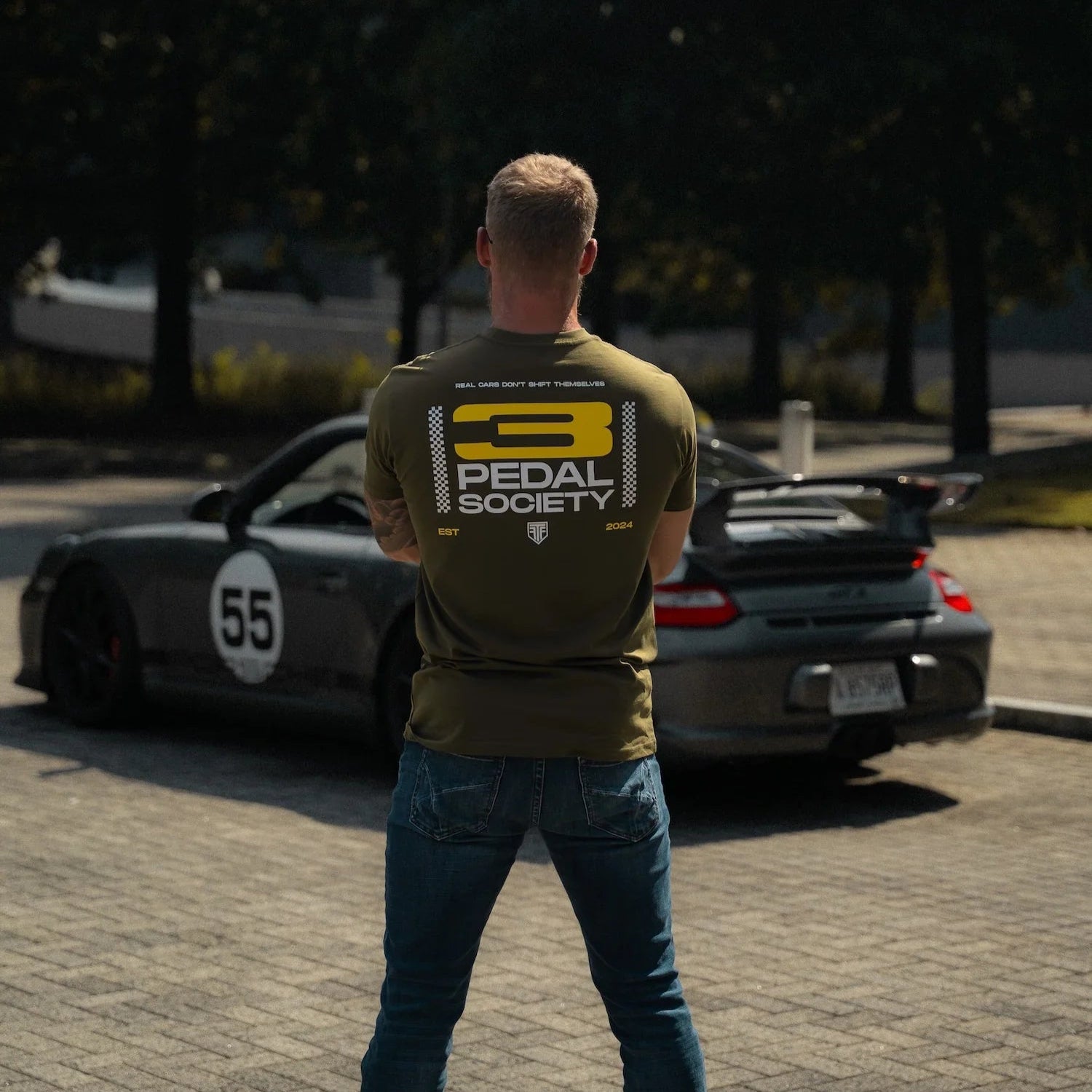Man wearing a green t-shirt with 'Pedal Society' logo standing in front of a black car with number 55.