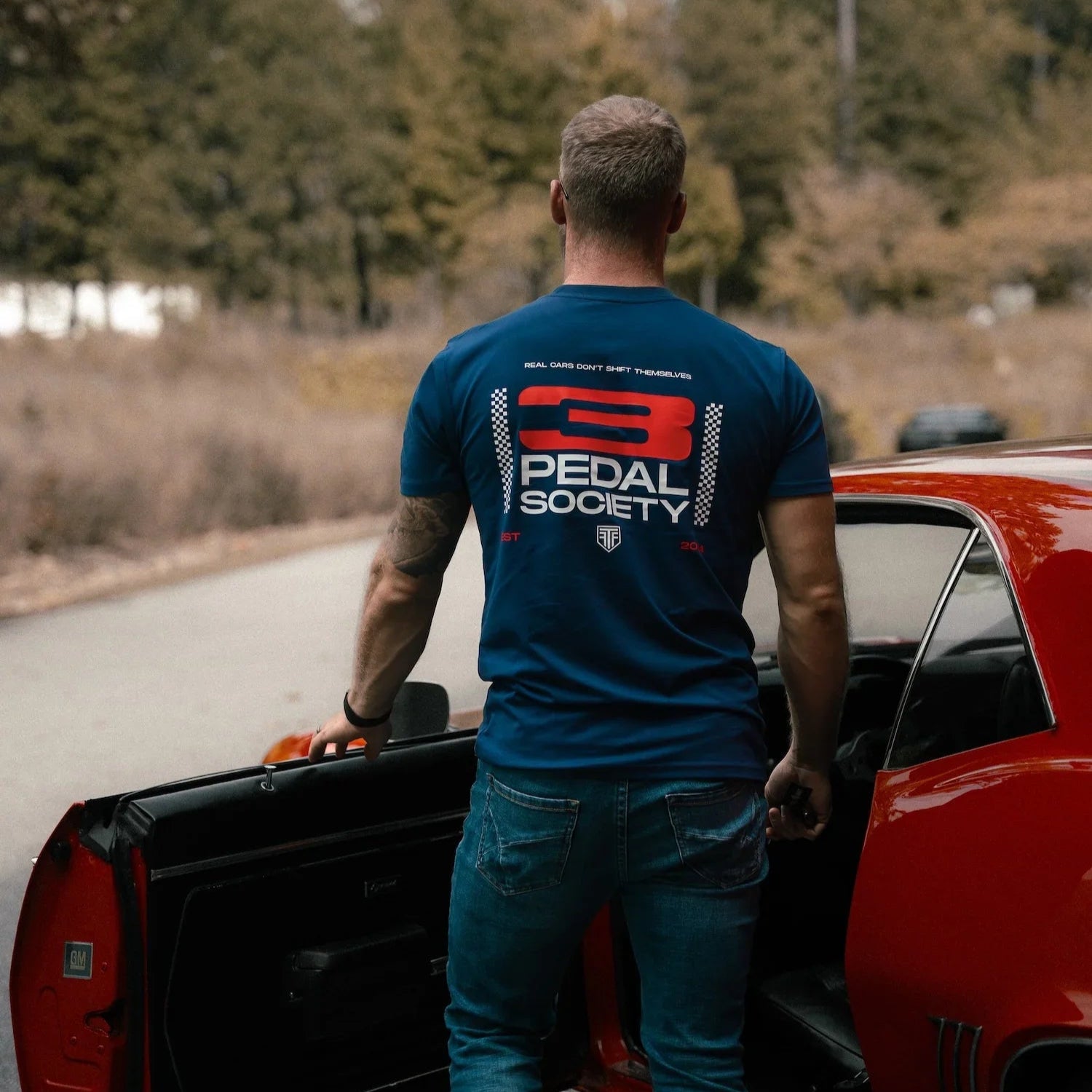 Man in a blue t-shirt with '3 Pedal Society' logo standing next to a red car on a road.