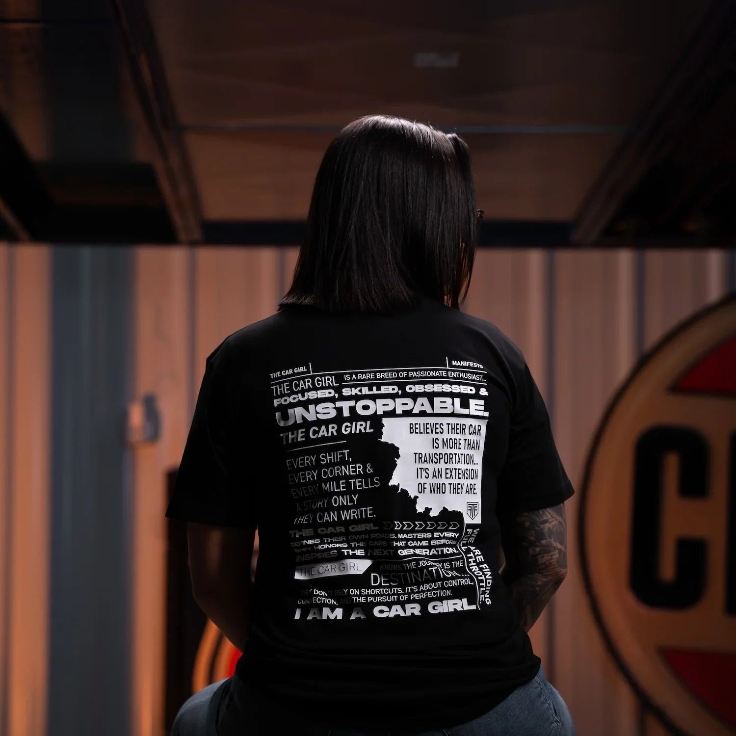 Person wearing a black t-shirt with text and graphics, sitting on a stool in a dimly lit room.