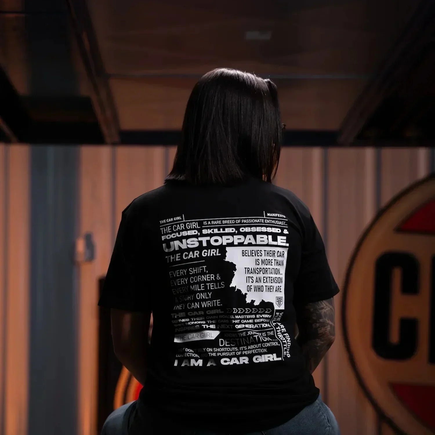 Person wearing a black t-shirt with text and graphics, sitting on a stool in a dimly lit room.