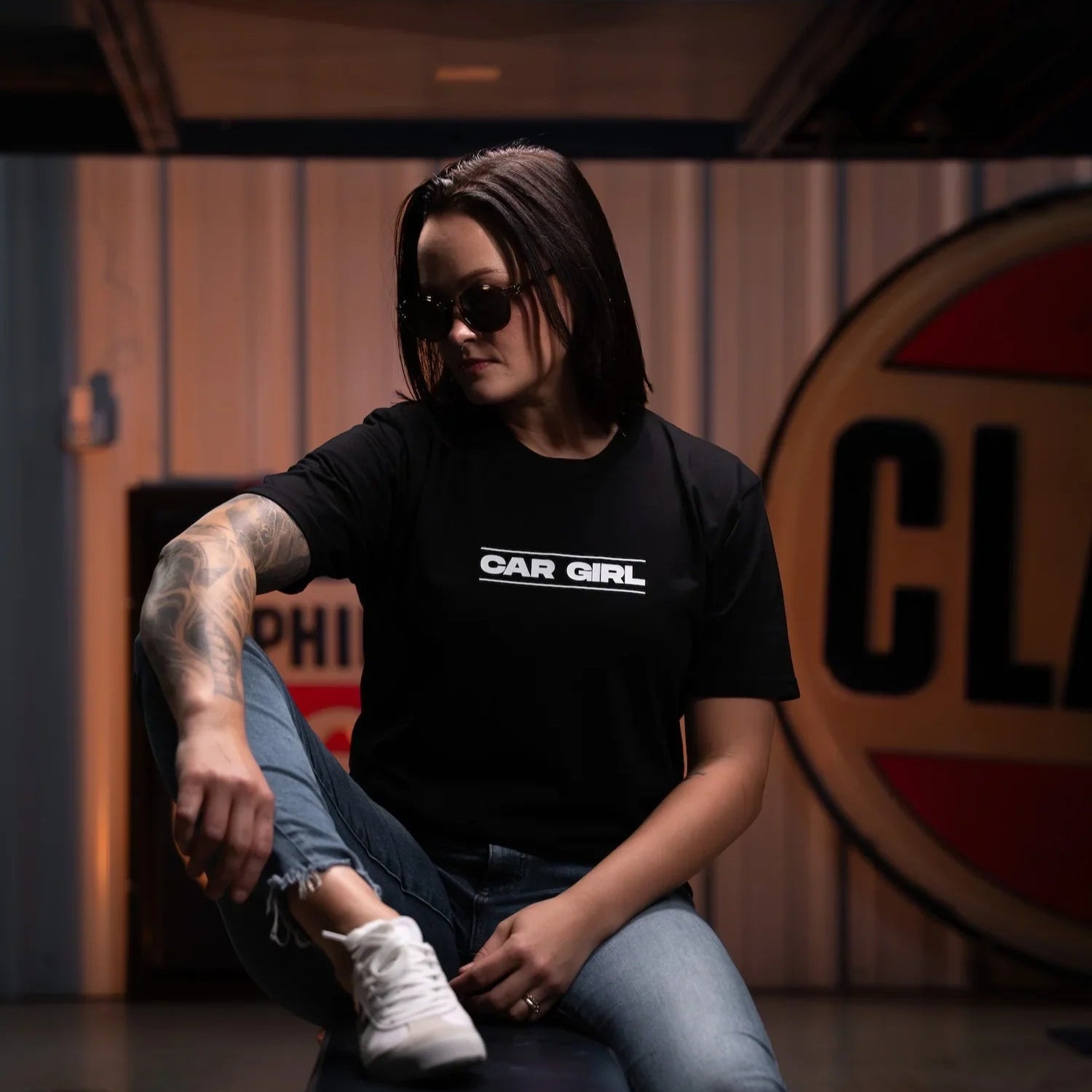Person wearing a black t-shirt with 'CAR GIRL' text, sitting on a bench in a garage setting.