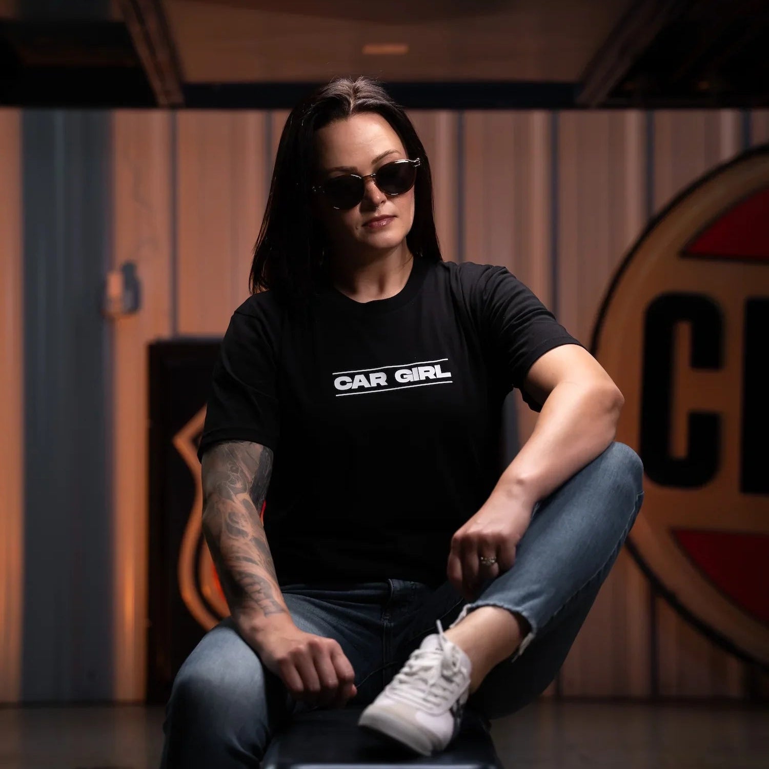 Person wearing a black t-shirt with 'CAR GIRL' text, sitting on a stool in a room with a logo on the wall.