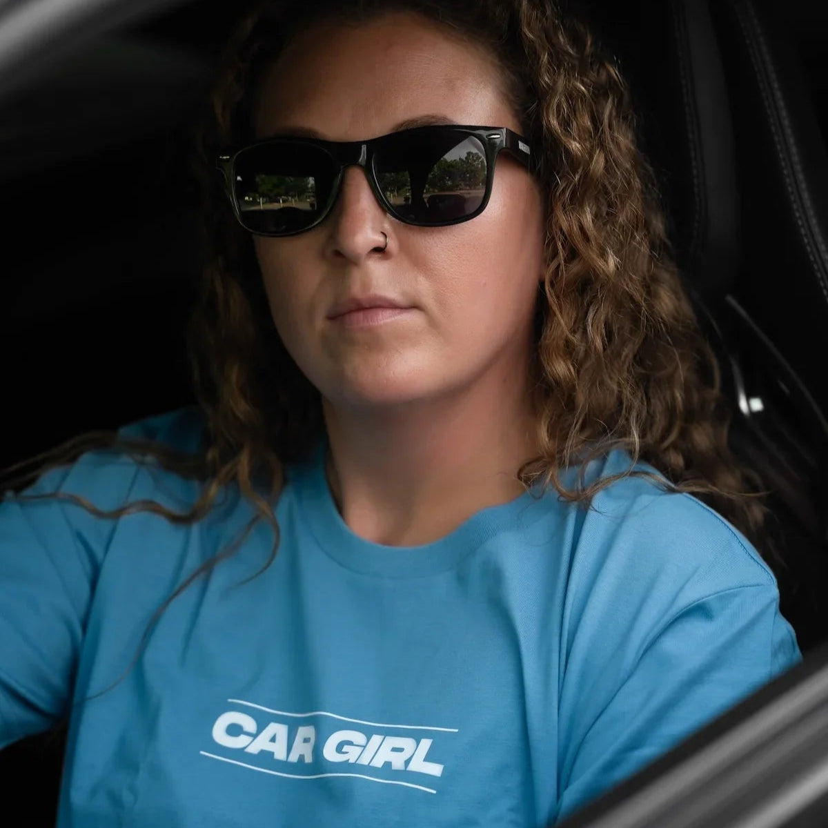 Woman wearing sunglasses and a blue 'CAR GIRL' t-shirt inside a car.