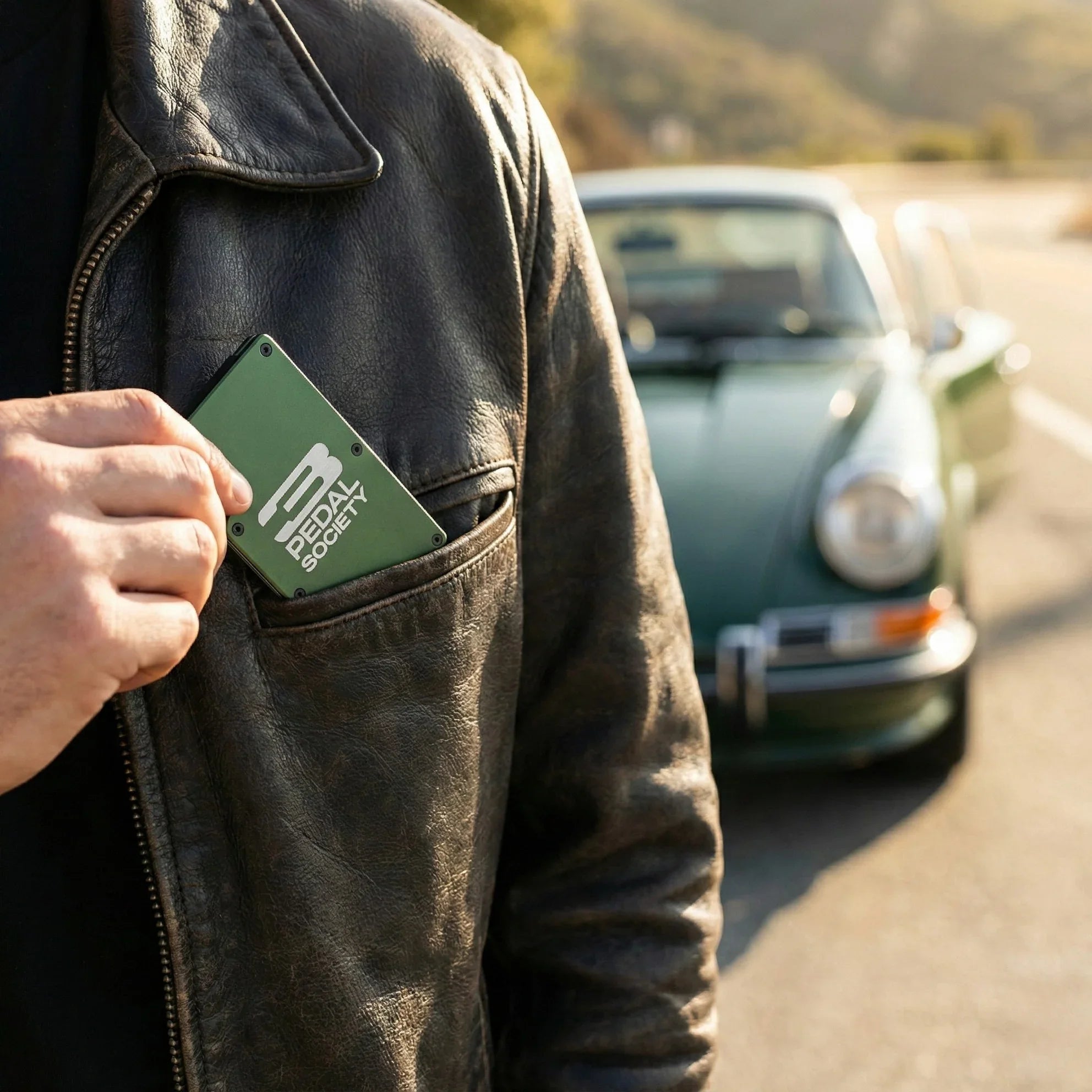 Person holding a green '3 Pedal Society' metal wallet with a logo in front of a vintage car on a road.