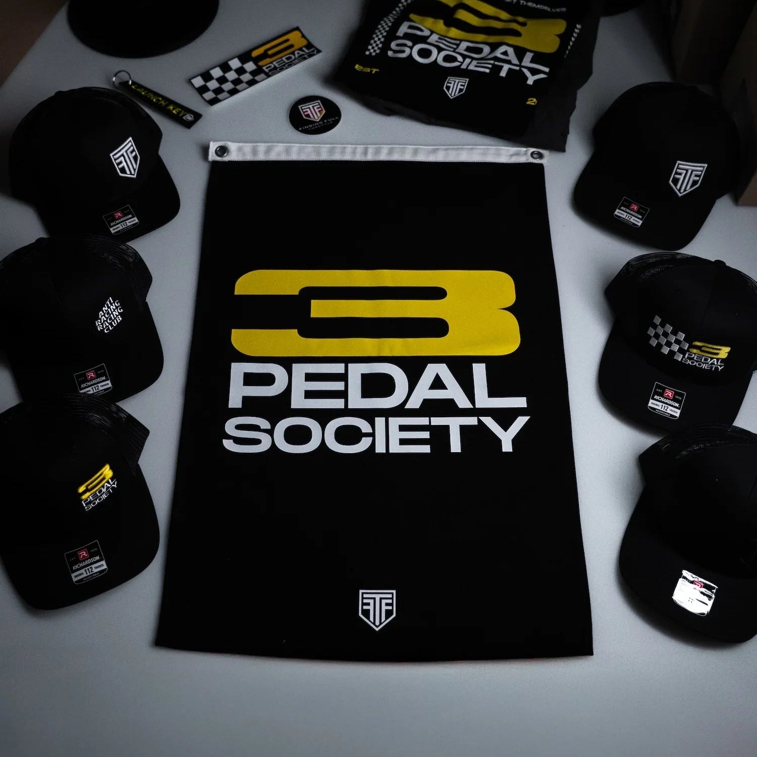Collection of '3 Pedal Society' merchandise including hats and a towel on a dark background.