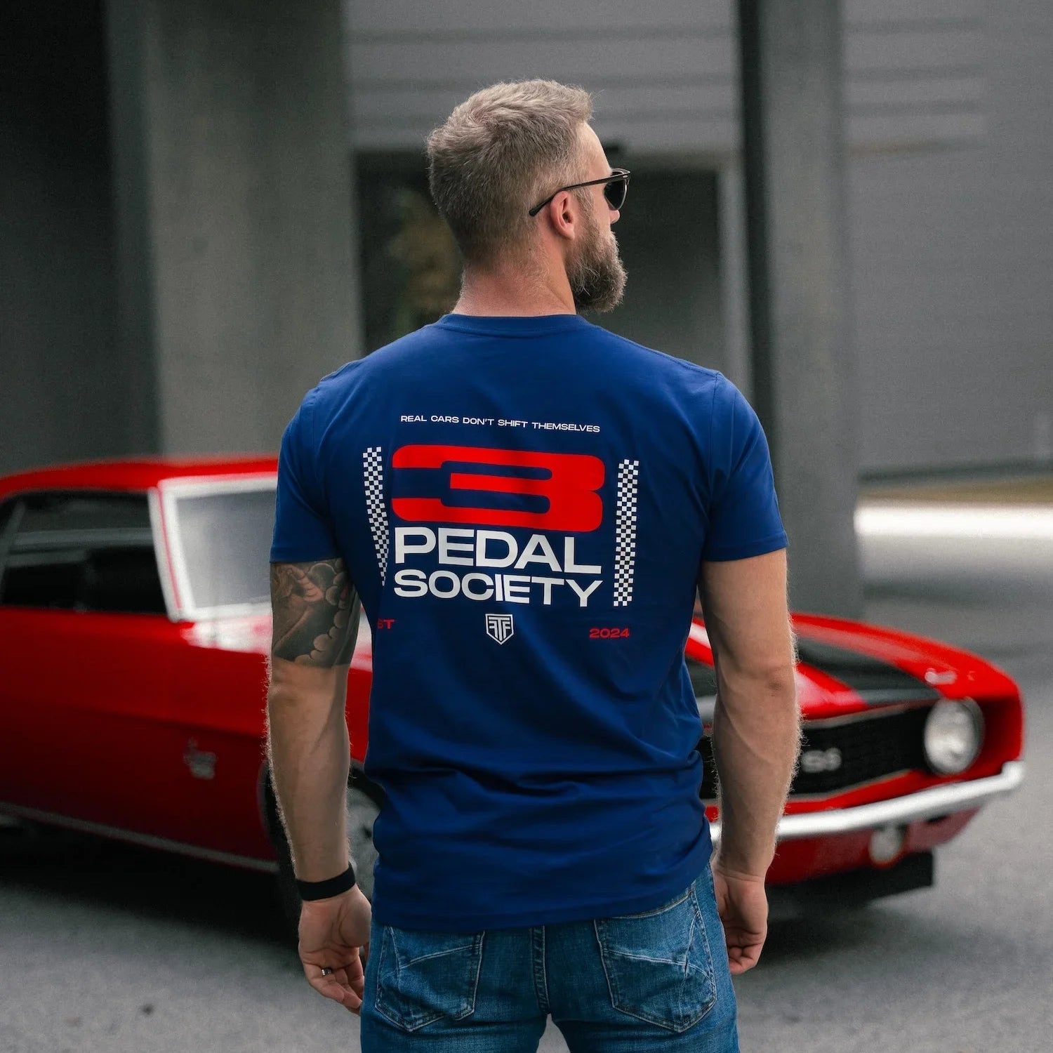 Man wearing a blue t-shirt with '3 Pedal Society' logo in front of a red car.