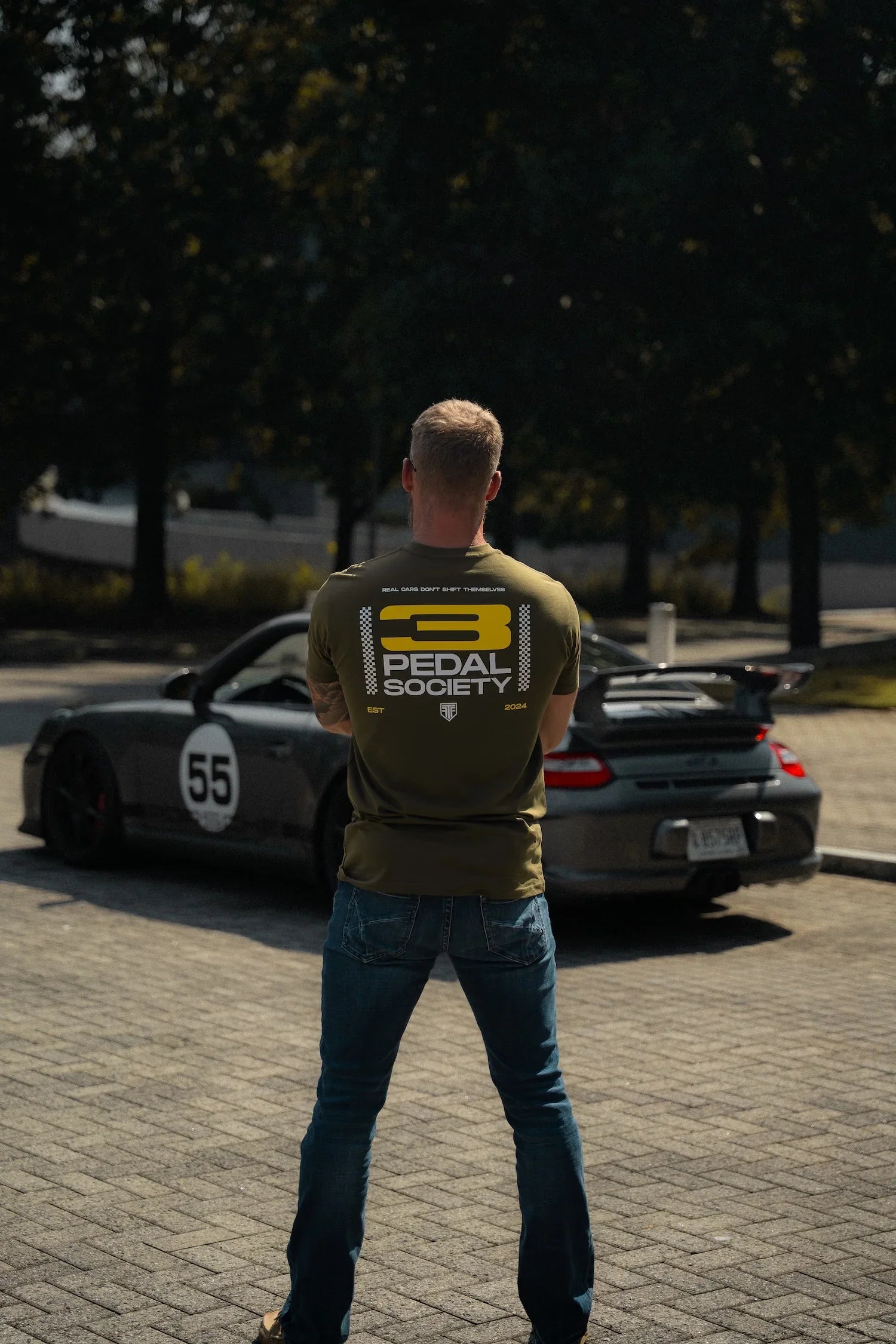 Man wearing a 'Pedal Society' shirt standing in front of a car with a visible license plate.