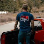 Man in a blue t-shirt with '3 Pedal Society' logo standing next to a red car on a road.