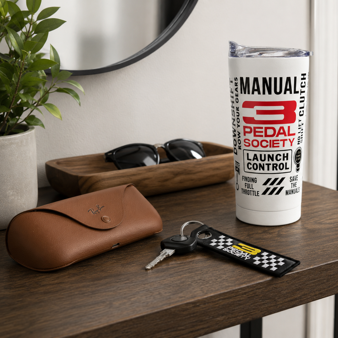 A white insulated tumbler with a clear lid stands on a wooden table, printed with the text and logo for 'Manual 3 Pedal Society'. The scene is styled as a lifestyle shot with a brown leather glasses case, sunglasses, keys, and a potted plant in the background.