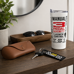 A white insulated tumbler with a clear lid stands on a wooden table, printed with the text and logo for 'Manual 3 Pedal Society'. The scene is styled as a lifestyle shot with a brown leather glasses case, sunglasses, keys, and a potted plant in the background.
