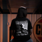Person wearing a black t-shirt with text and graphics, sitting on a stool in a dimly lit room.