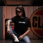 Person wearing a black t-shirt with 'CAR GIRL' text, sitting on a bench in a garage setting.