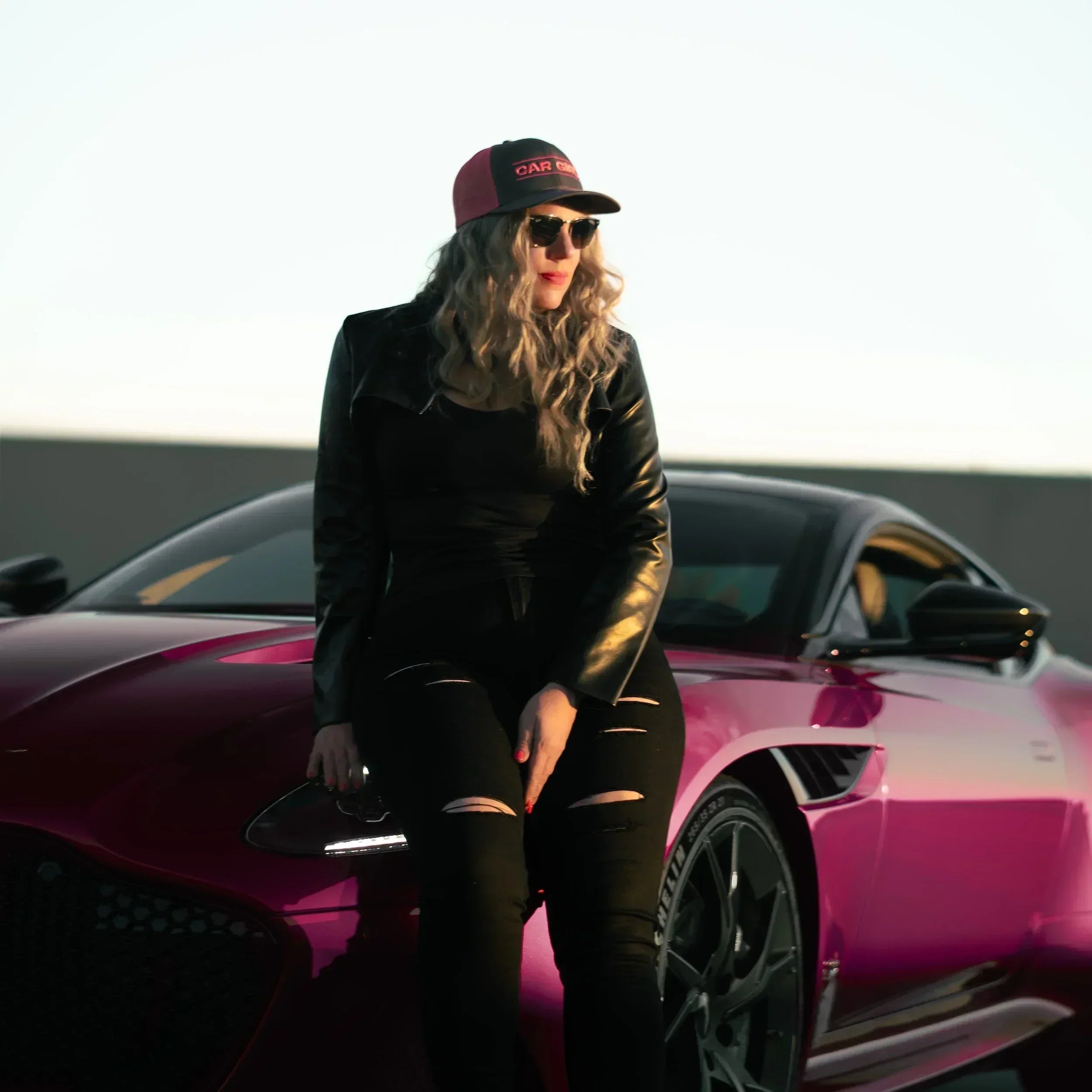 Person wearing a black outfit and red cap sitting on a pink sports car with a clear sky background