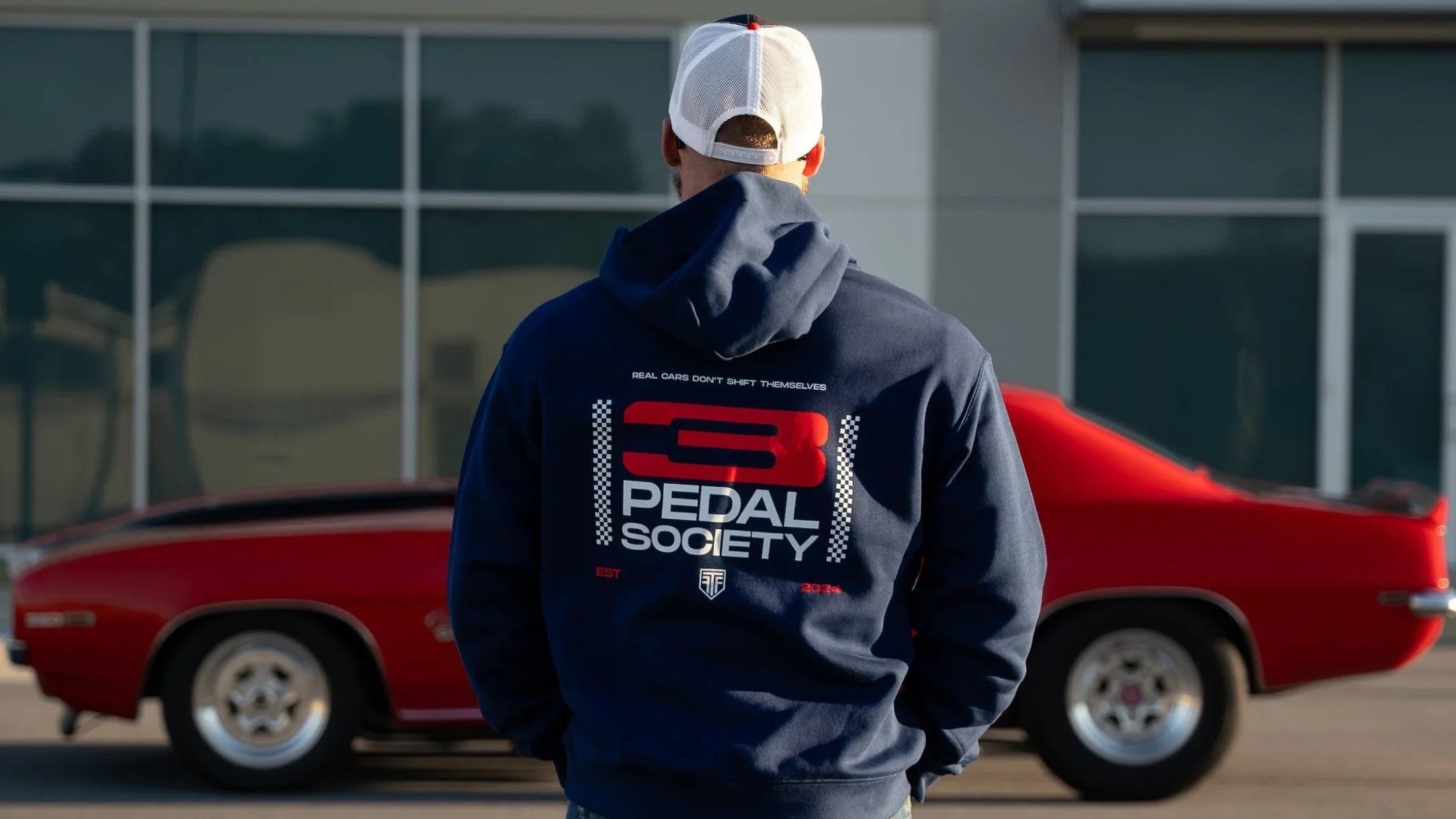 Person wearing a hoodie with 'Pedal Society' logo in front of a red car.