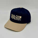 A two-tone baseball cap with a navy blue crown and a beige curved brim, featuring a white embroidered '3 PEDAL SOCIETY' logo with a checkered flag design on the front.