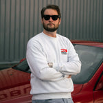 Man wearing sunglasses and a white sweatshirt with a logo, standing in front of a red car.