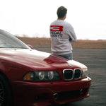 Person wearing a white t-shirt with a logo, standing next to a red car on a road.