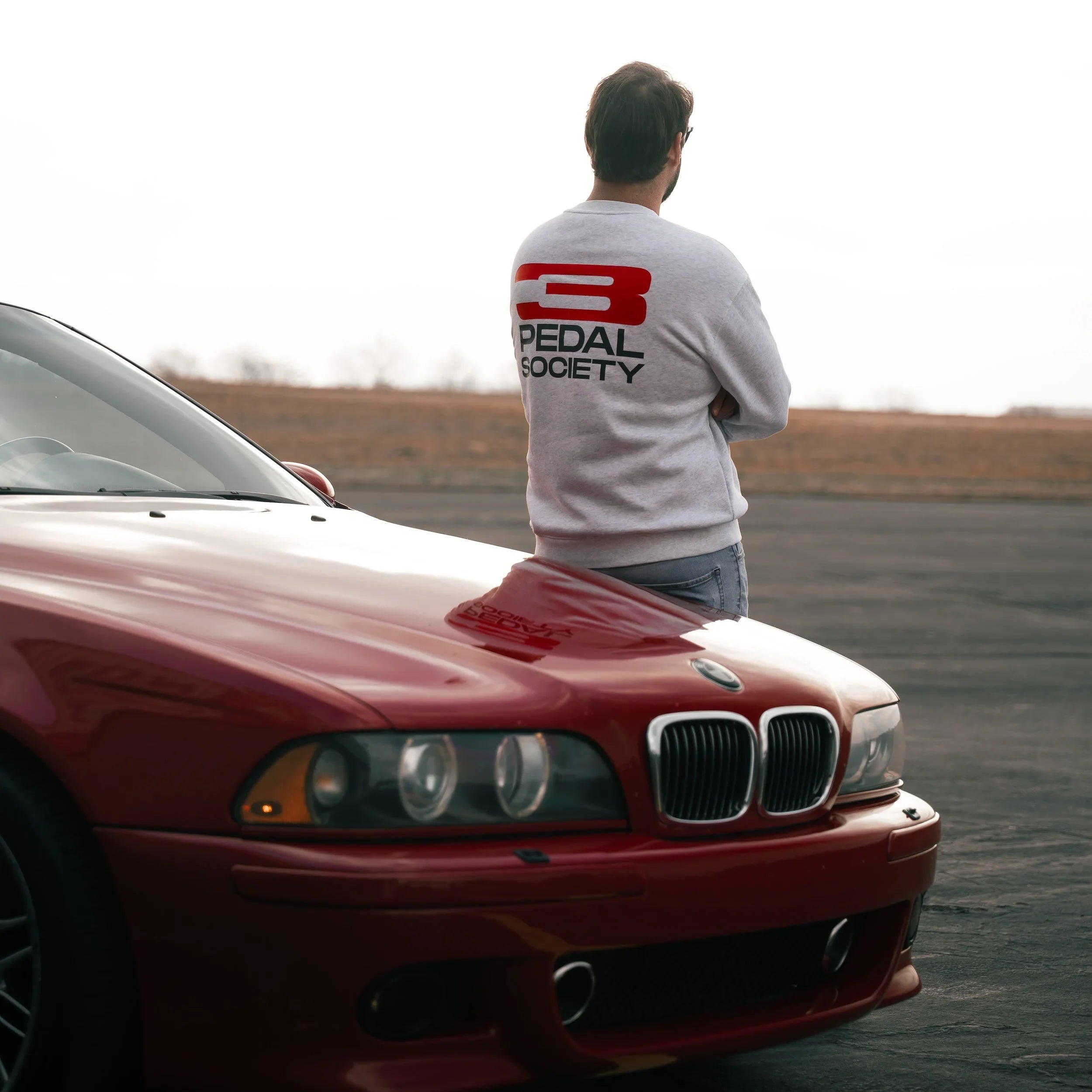 Person wearing a white t-shirt with a logo, standing next to a red car on a road.