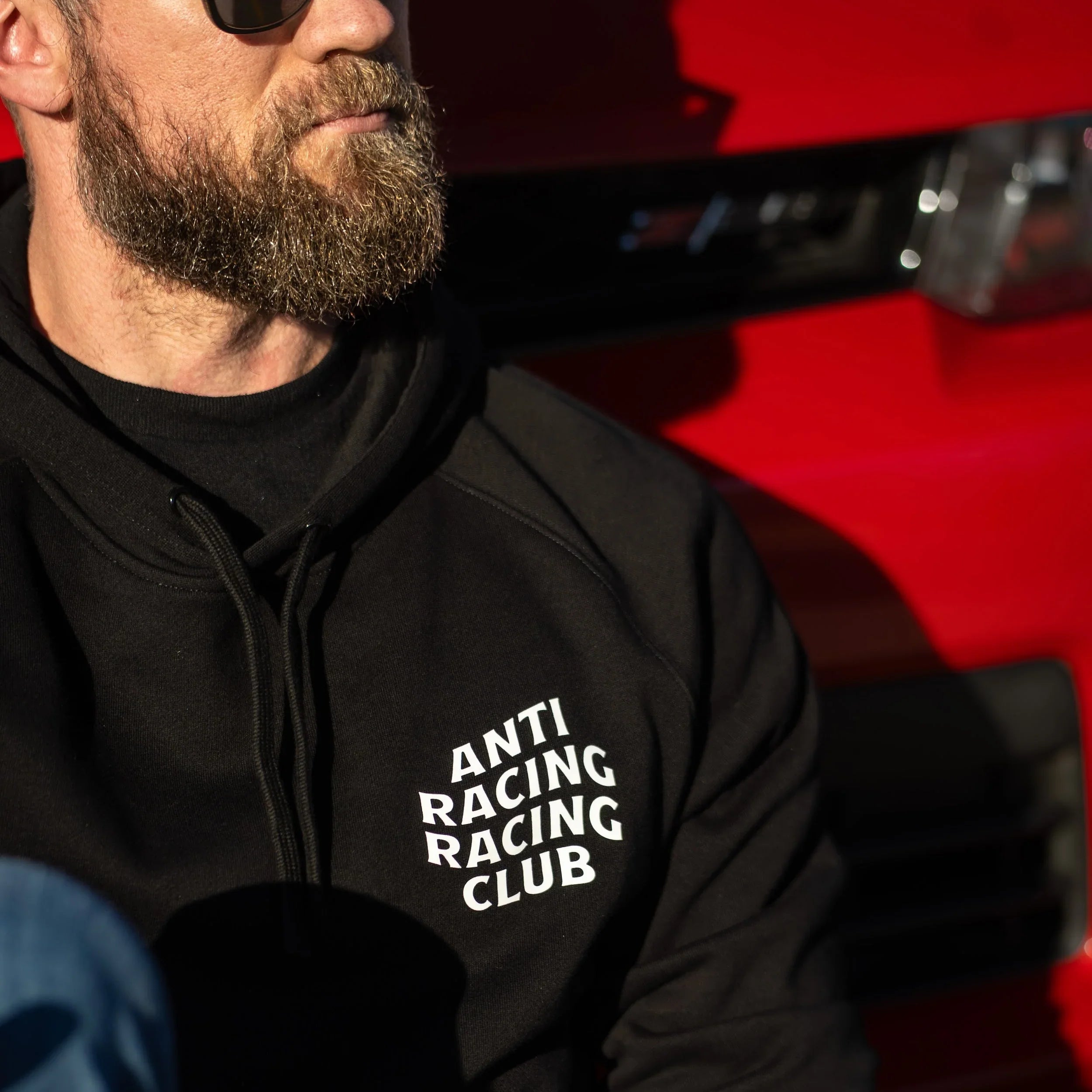Person wearing a black hoodie with 'ANTI RACING RACING CLUB' text, sitting in front of a red vehicle.