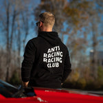 Person wearing a black hoodie with 'ANTI RACING RACING CLUB' text, standing by a red car with trees in the background.