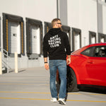 Man wearing a black hoodie with 'ANTI RACING CLUB' text, walking towards a red car in an industrial area.