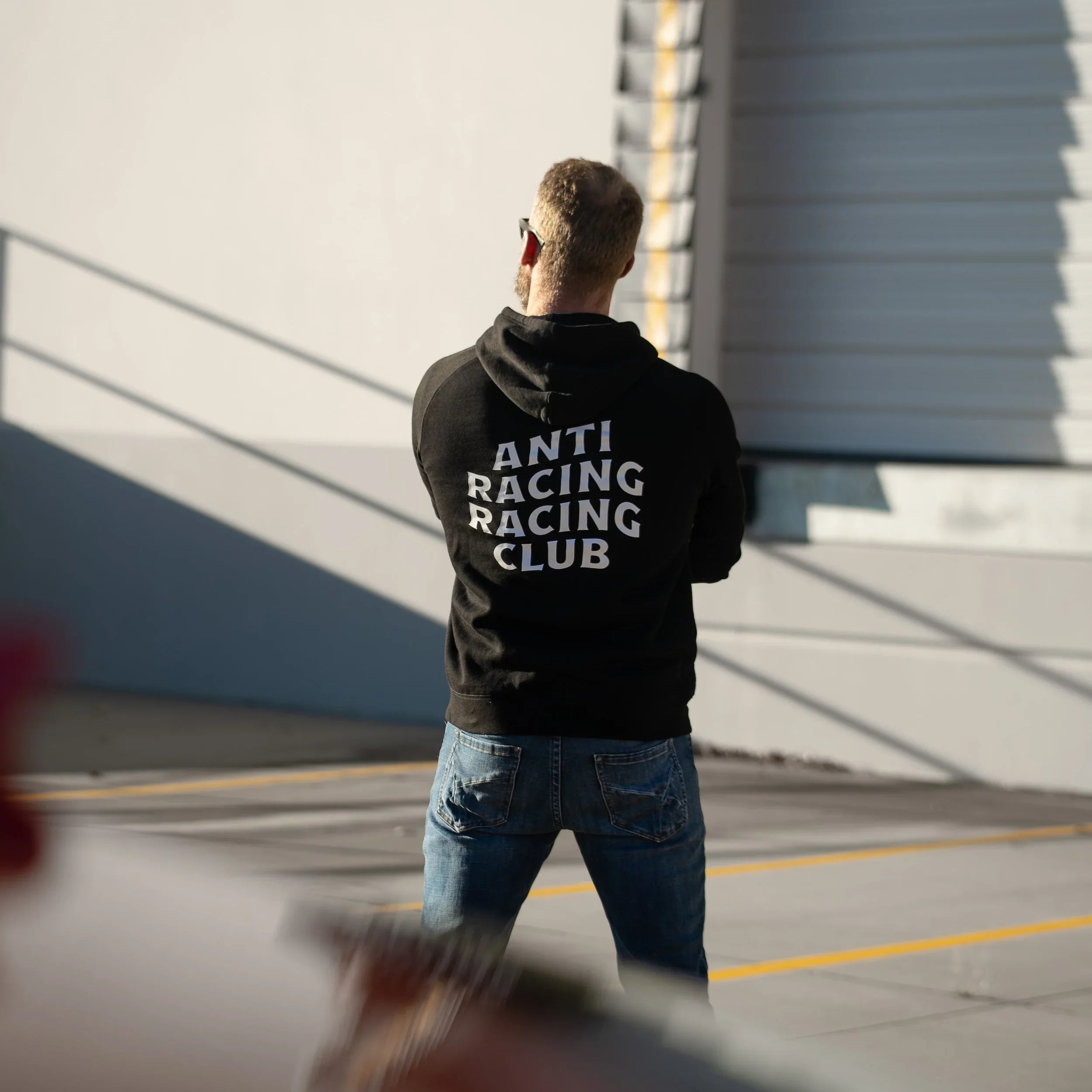 Person wearing a black hoodie with 'ANTI RACING CLUB' text, standing in front of a building.