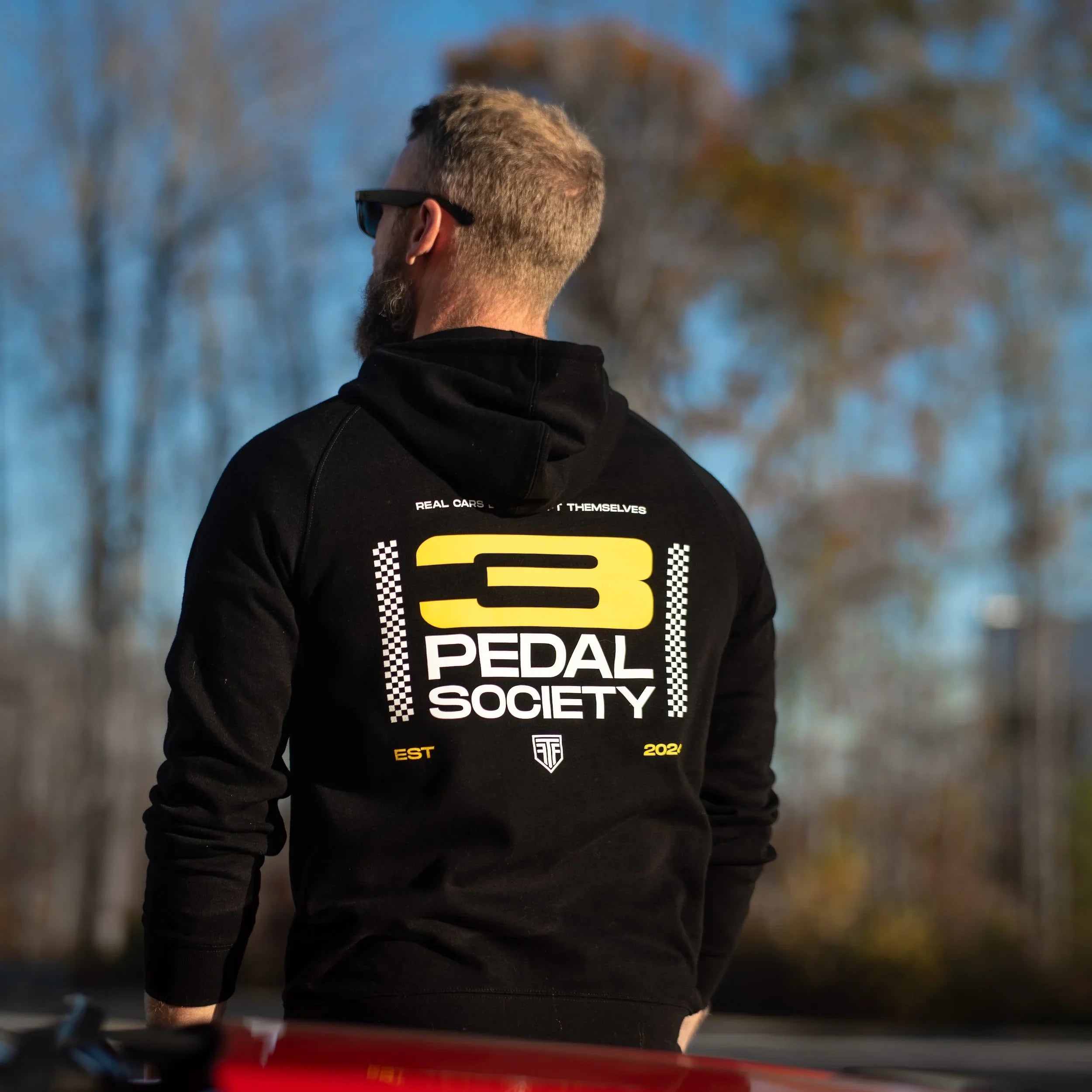 Person wearing a black hoodie with '3 Pedal Society' logo, standing in front of a red vehicle with trees in the background.