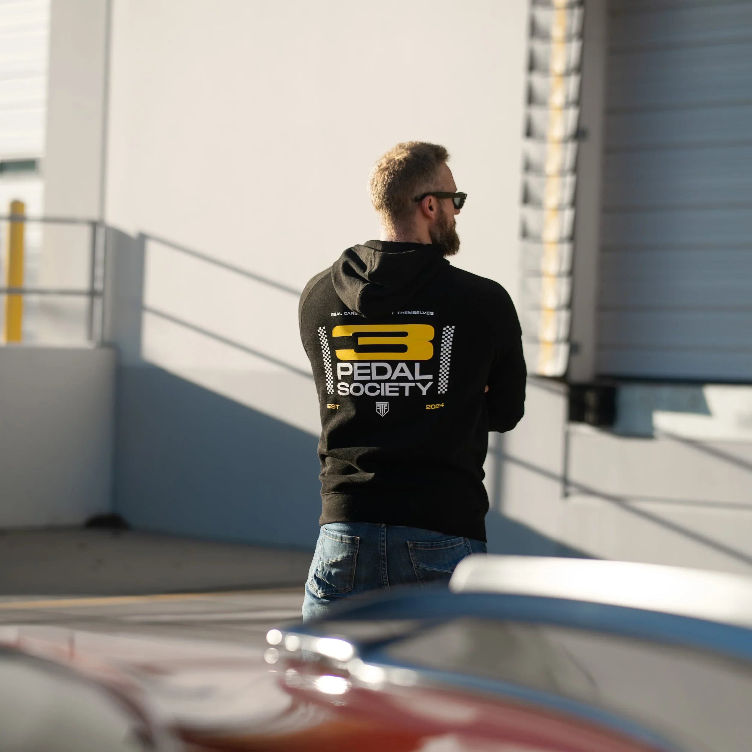 Person wearing a '3 Pedal Society' hoodie standing in front of a building with a car in the foreground.