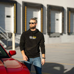 Man standing next to a red car wearing a black hoodie with a logo, in front of a building with open garage doors.