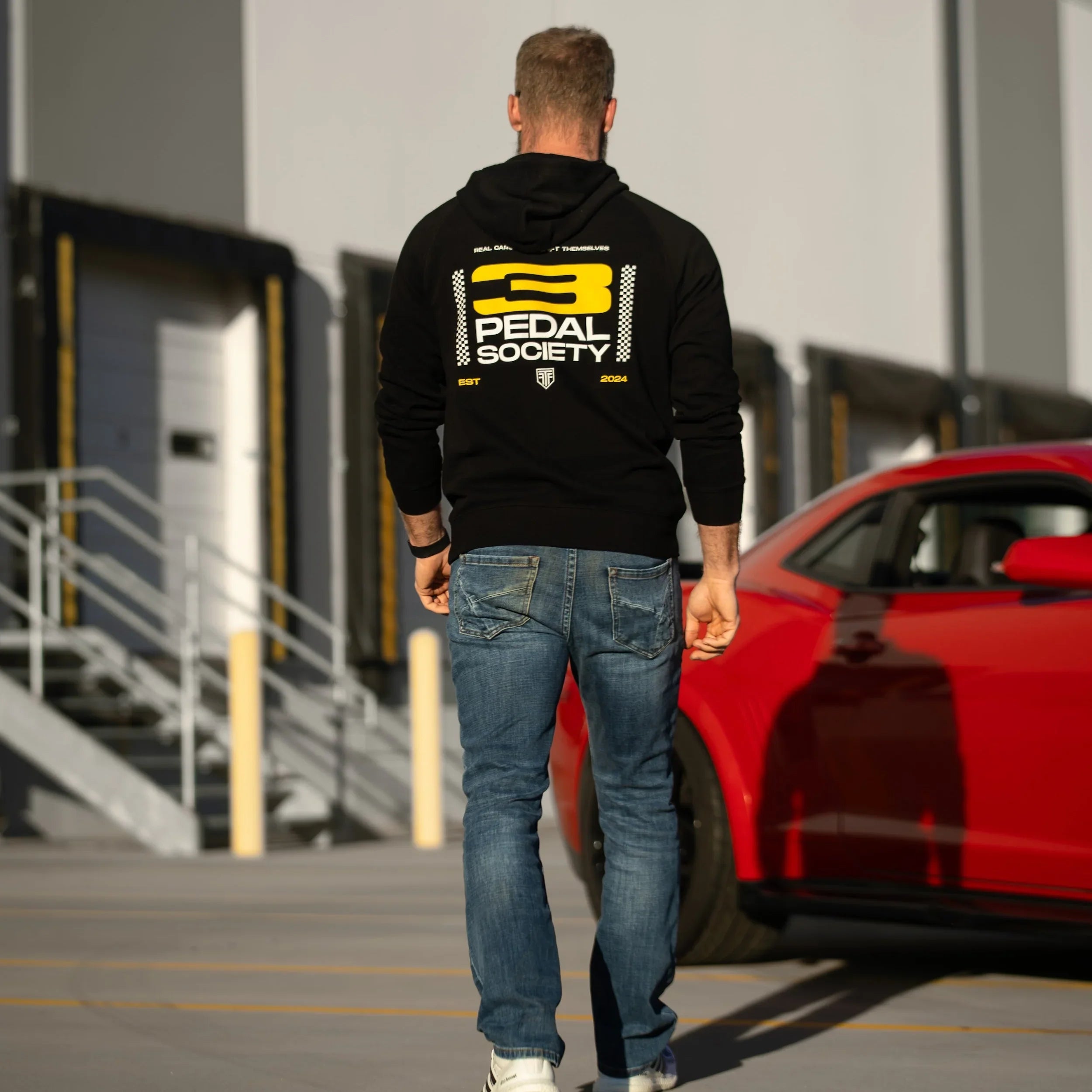 Person wearing a black hoodie with a logo, walking towards a red car in front of a building.