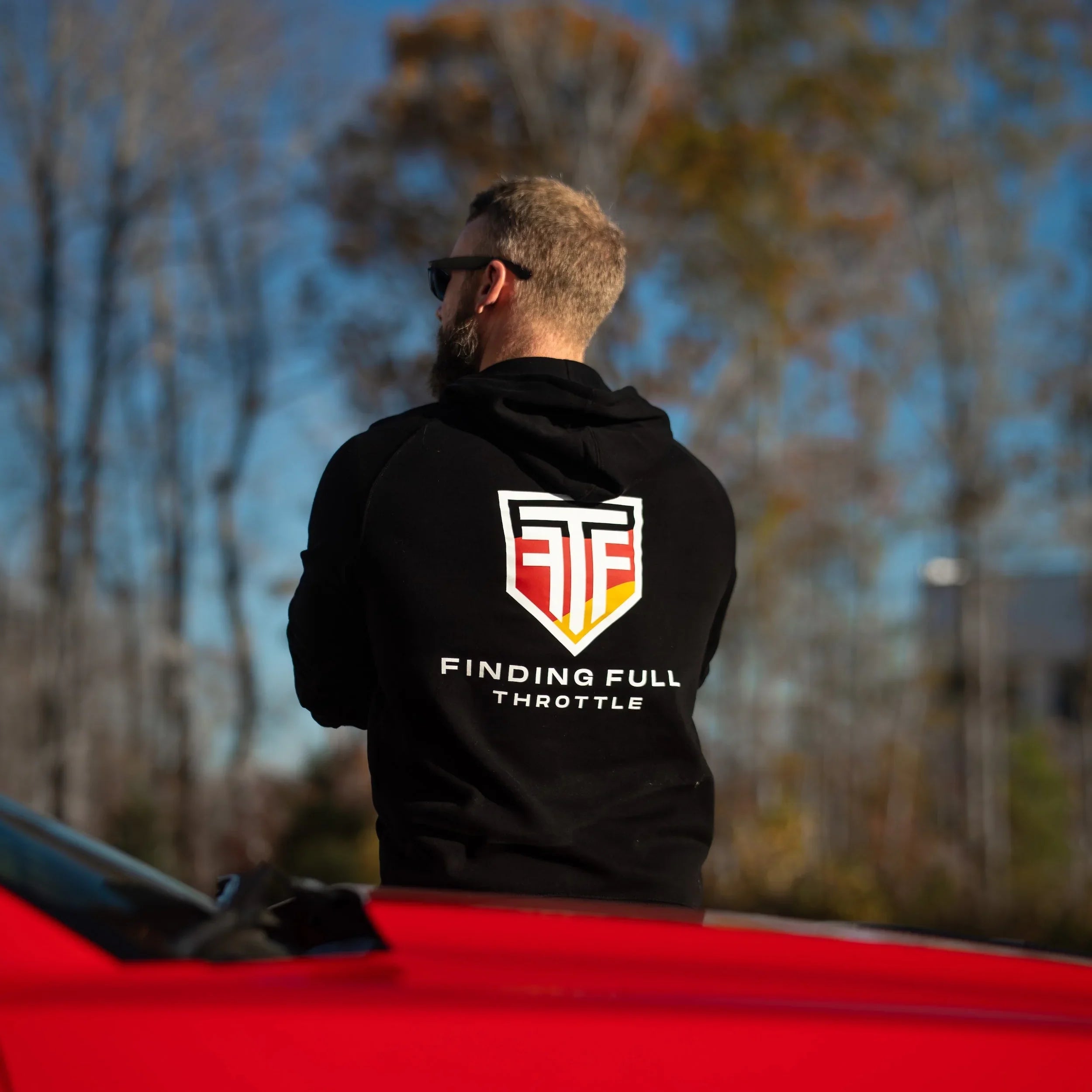 Person wearing a black hoodie with a logo and text, sitting on a red car with trees and blue sky in the background.