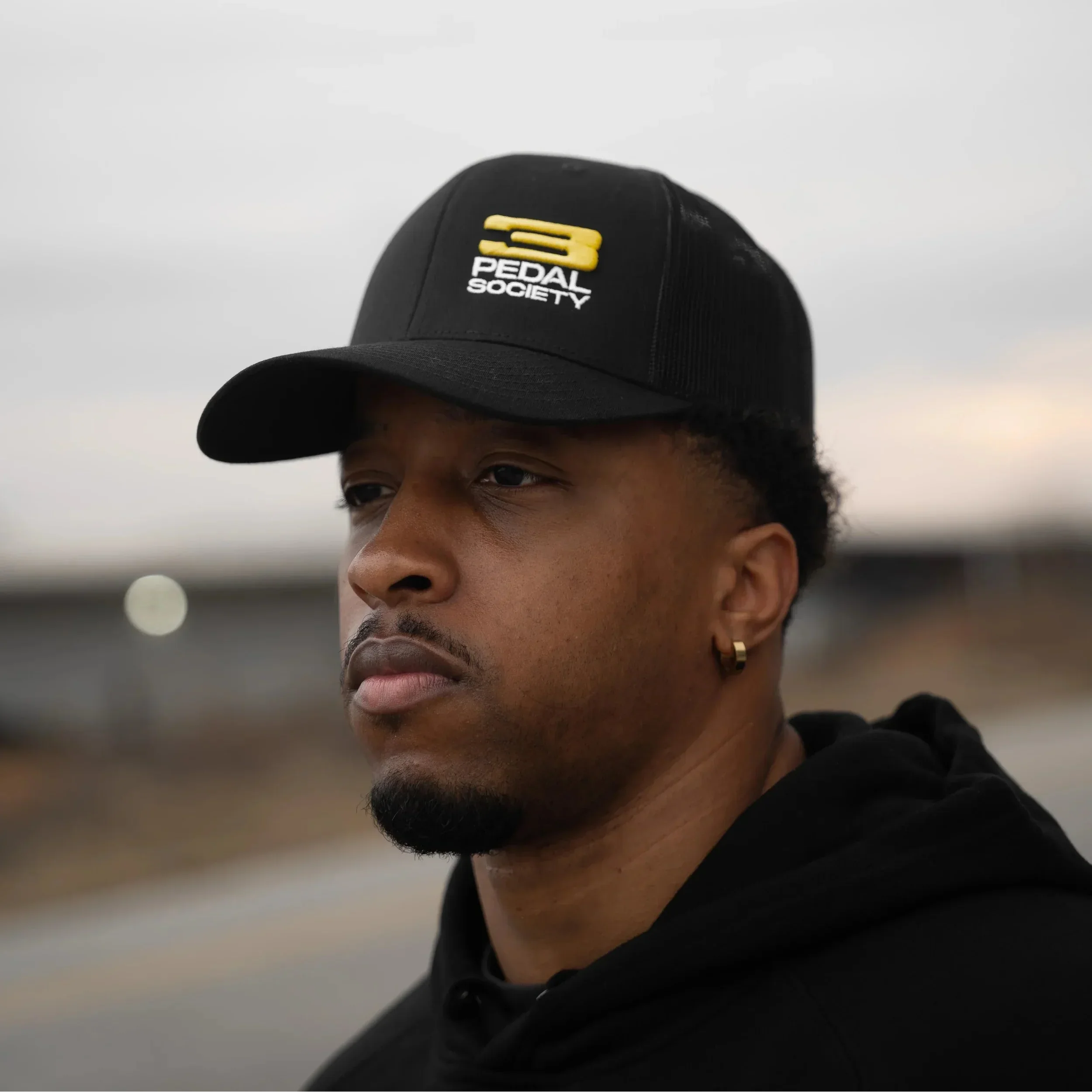 Man wearing a black 3 Pedal Society hat with a logo against a blurred background