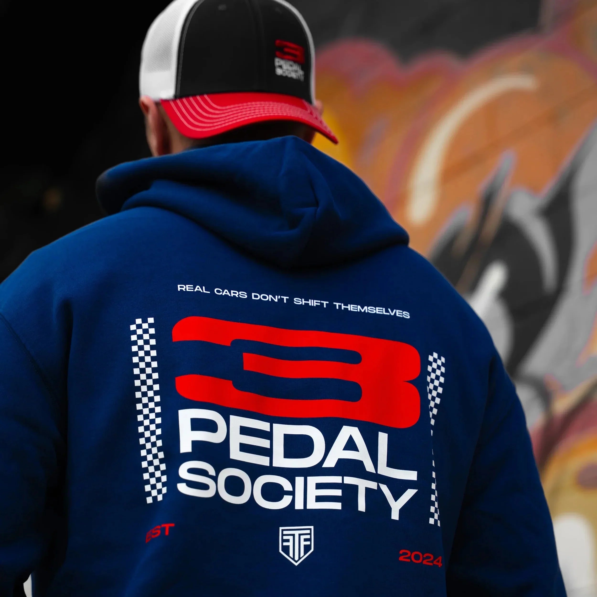 Person wearing a blue hoodie with '3 Pedal Society' branding in front of graffiti art.