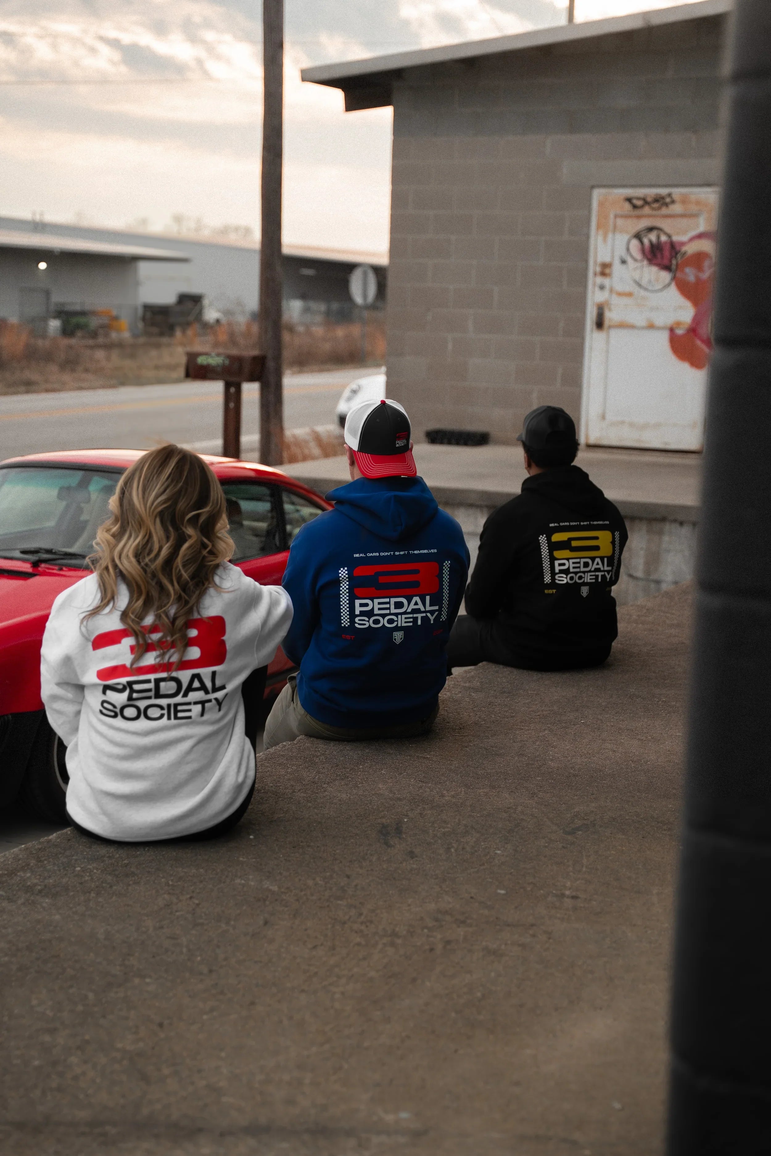 Three people sitting on a concrete surface with '3 pedal society ' branded clothing, facing a red car.