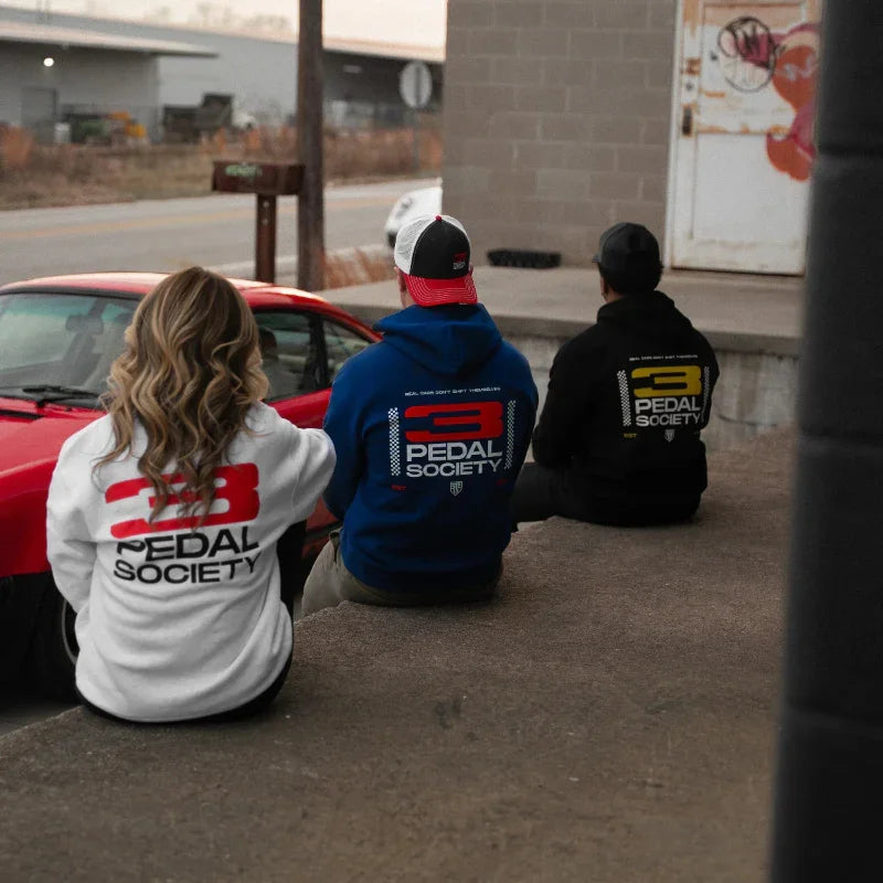 Three people sitting on a curb wearing hoodies with 'Pedal Society' branding, in front of a red car.