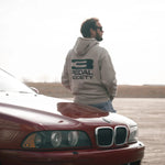 A man wearing a light gray hoodie with a black '3 PEDAL SOCIETY' logo on the back stands next to a red classic car in an open outdoor setting.