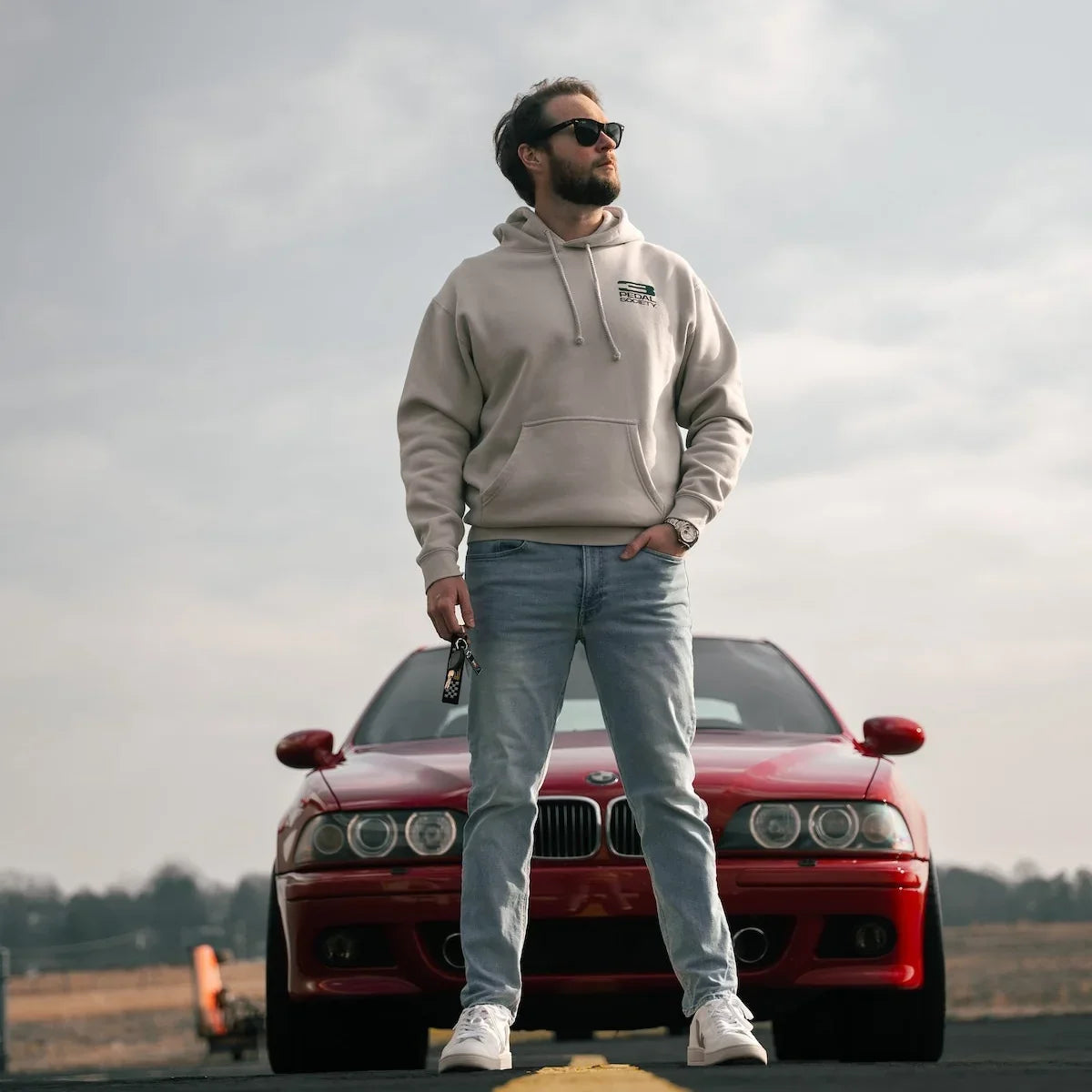 A man wearing a beige hoodie, light blue jeans, and white sneakers stands on an airstreet in front of a red BMW M3 sports car, looking off-camera under a cloudy sky.
