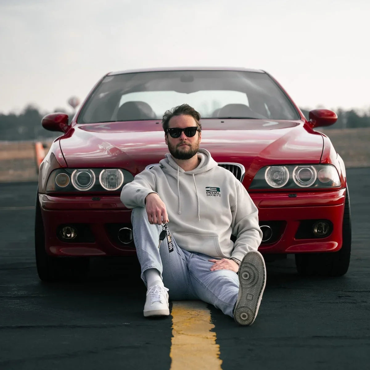 A man wearing a light gray hoodie, light gray jeans, and white sneakers sits on the front bumper of a red BMW M5 on an asphalt road with a yellow line.