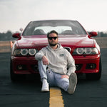 A man wearing a light gray hoodie, light gray jeans, and white sneakers sits on the front bumper of a red BMW M5 on an asphalt road with a yellow line.