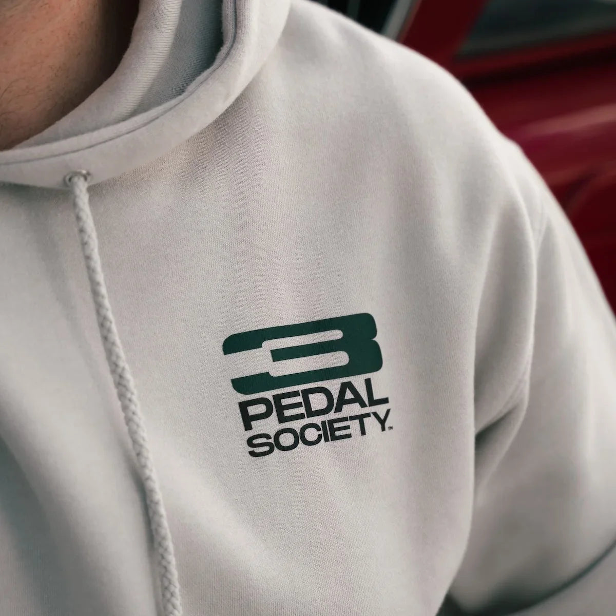 A close-up of a person wearing a light gray hoodie with a dark green '3 PEDAL SOCIETY.' logo printed on the left chest.