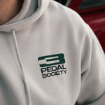A close-up of a person wearing a light gray hoodie with a dark green '3 PEDAL SOCIETY.' logo printed on the left chest.