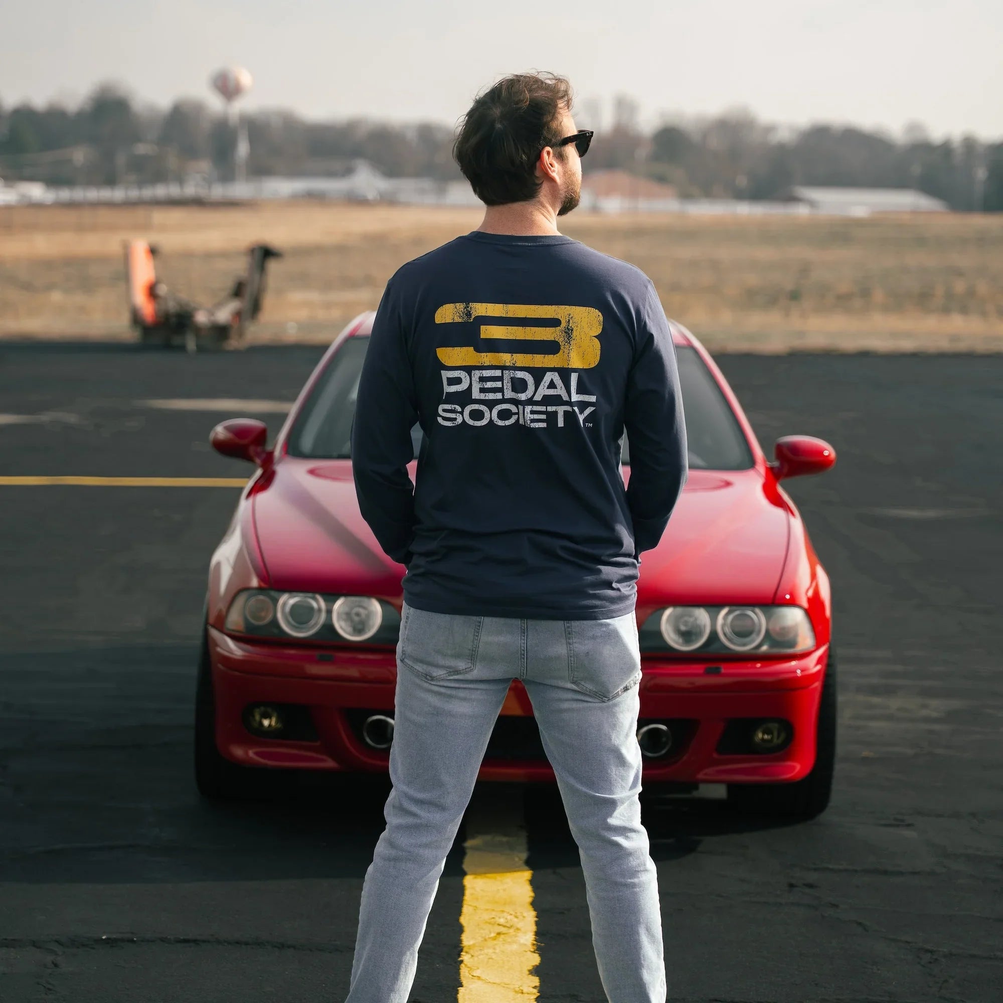 Person wearing a '3 Pedal Society' shirt standing in front of a red car on a runway.