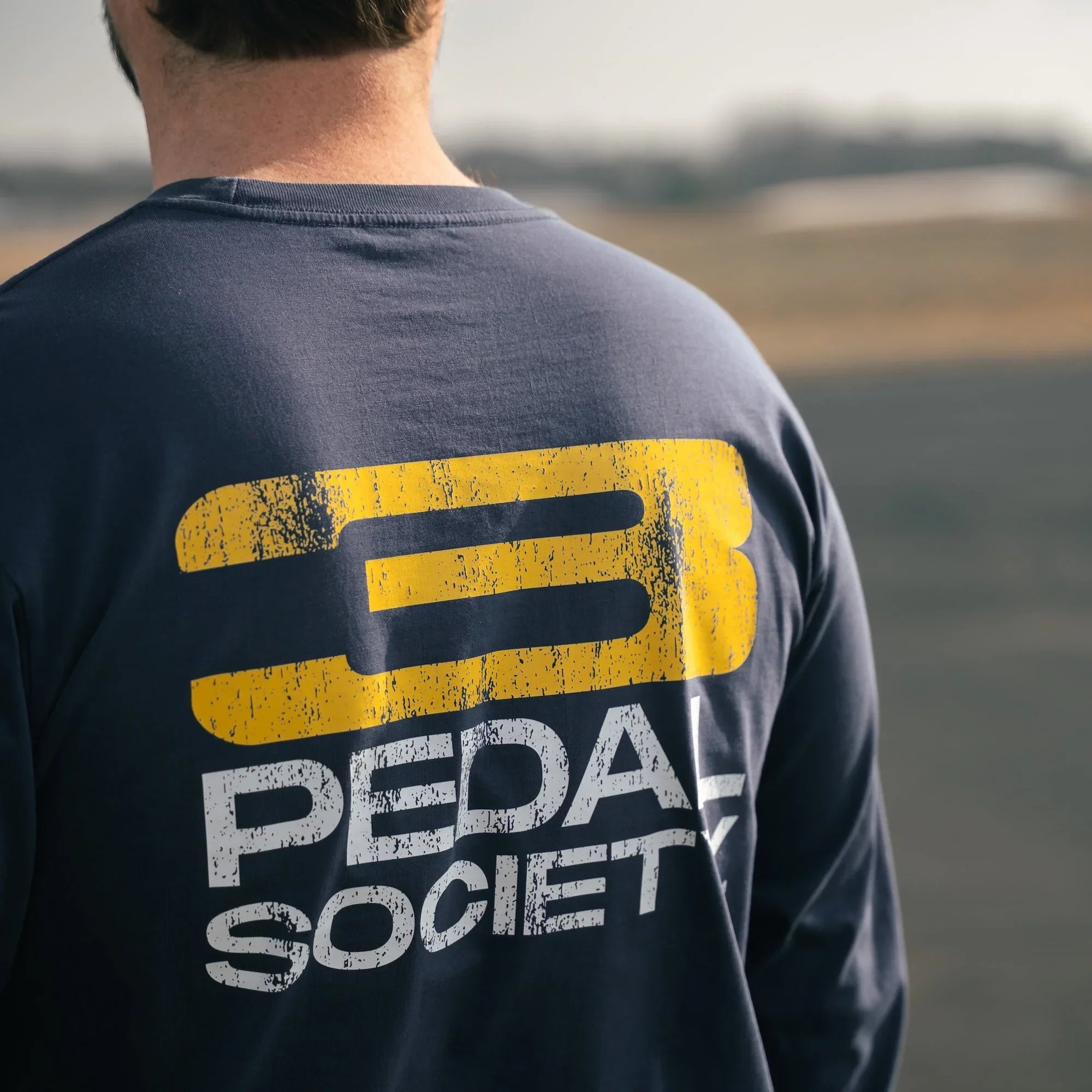 Person wearing a navy blue sweatshirt with 'Pedal Society' logo on the back, standing outdoors.