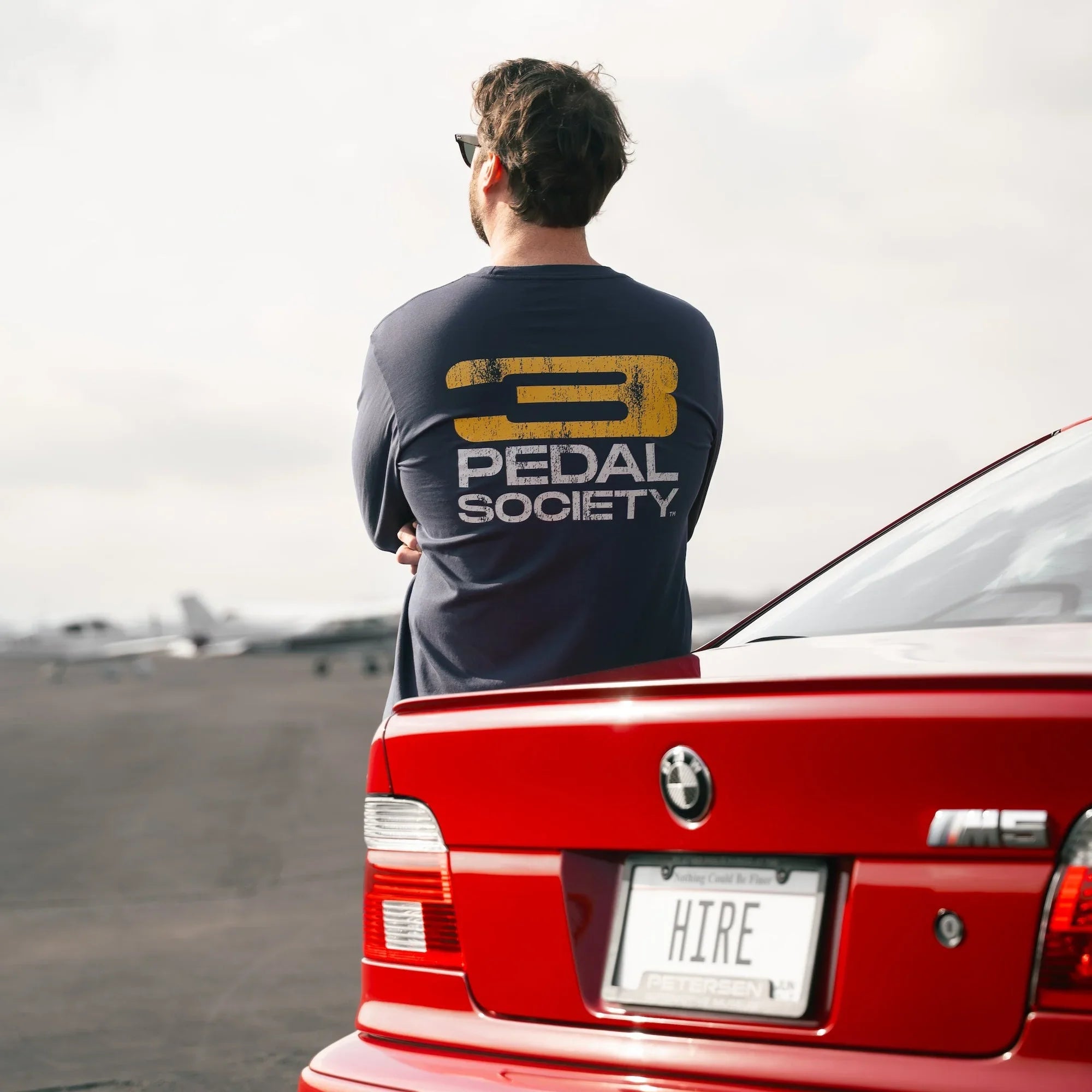 Person wearing a '3 Pedal Society' shirt standing next to a red BMW car with a cloudy sky.