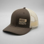 A two-tone trucker cap with a dark brown front panel and brim, and a beige mesh back. The front panel features a metallic gold embroidered logo and the text 'PEDAL SOCIETY'.