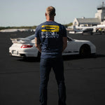 Person wearing a 'Pedal Society' t-shirt standing on a runway with a white car.