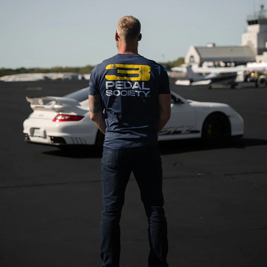 Person wearing a 'Pedal Society' t-shirt standing on a runway with a white car.