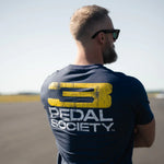 Man wearing a 'Pedal Society' t-shirt with a yellow and white logo on a clear day.