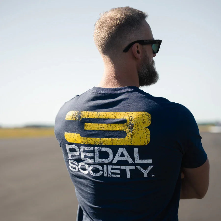 Man wearing a 'Pedal Society' t-shirt with a yellow and white logo on a clear day.